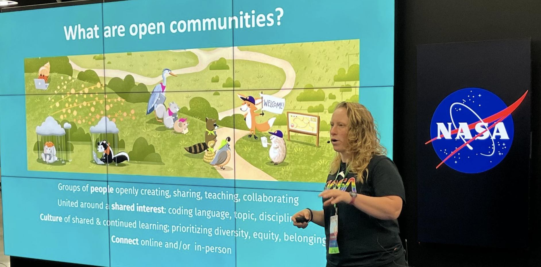 Photo of a white woman presenting, standing in front of a big NASA logo and a screen that says Open Communities with an illustration of friendly animals.