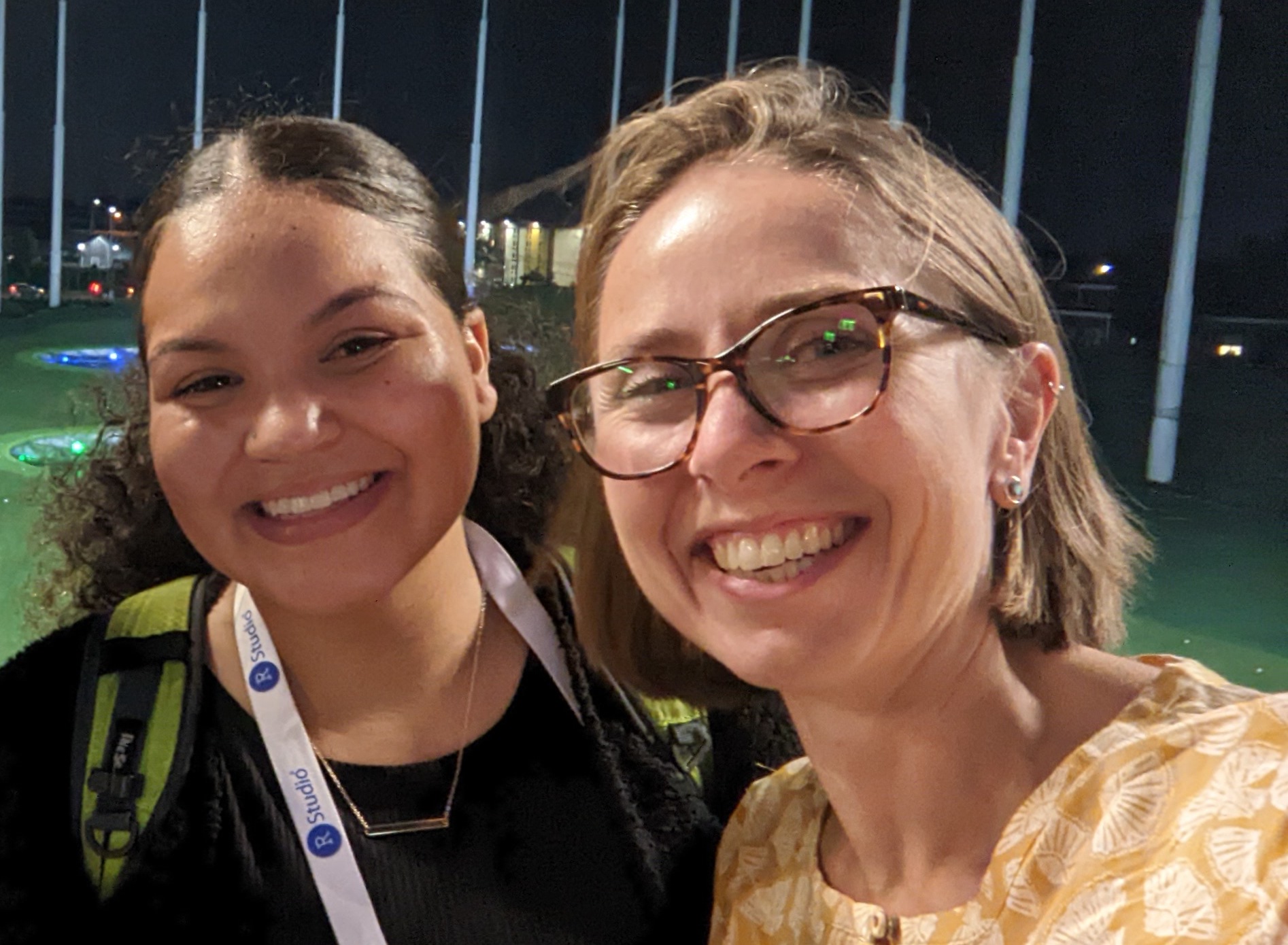 Two women smiling at the camera outside at night. The woman on the left is Black and wearing a black shirt and a lanyard that says RStudio. The woman on the right is white and wearing a yellow shirt and glasses.