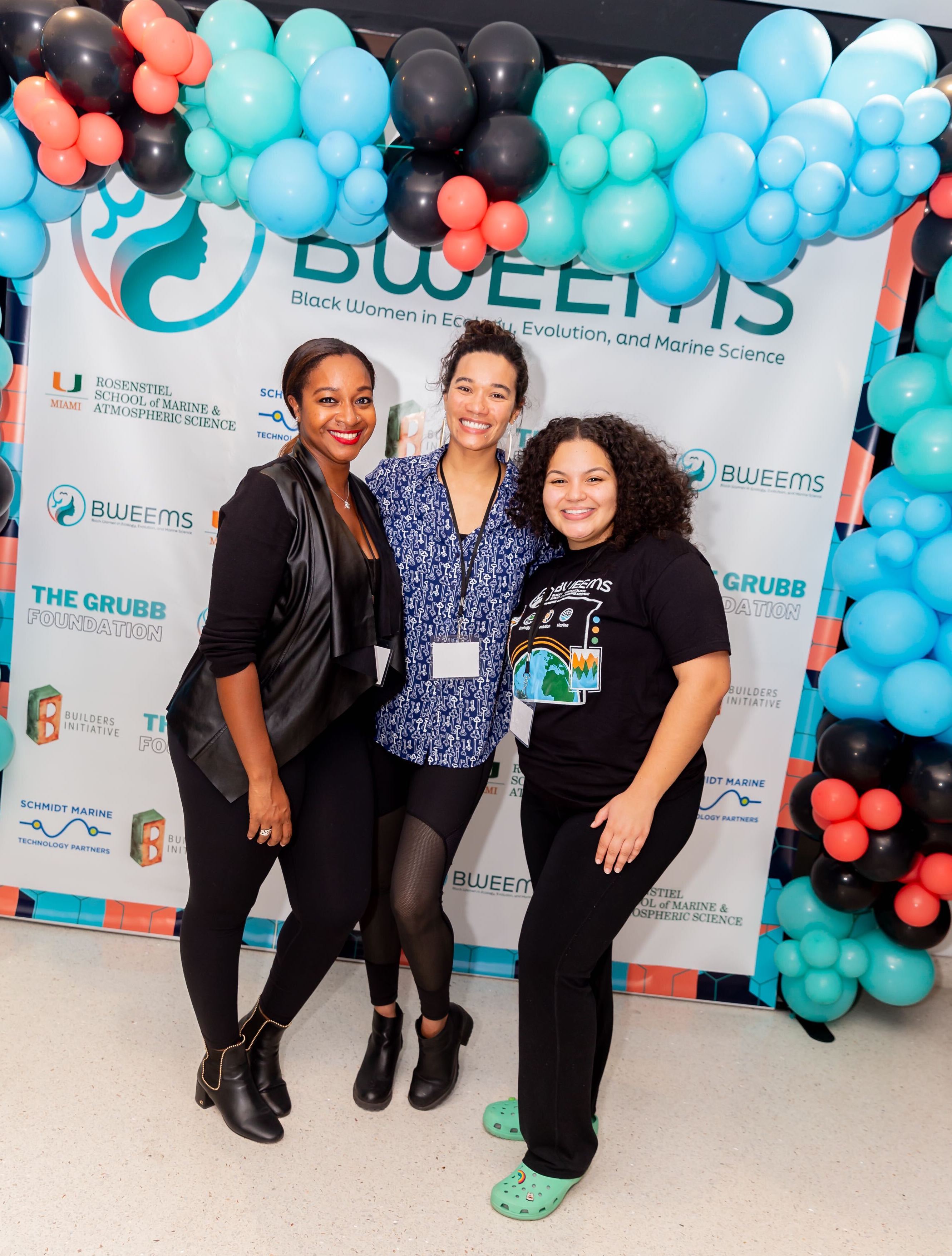 3 smiling Black women standing in front of a BWEEMS-branded backdrop with an arbor of black, blue, green, and red balloons