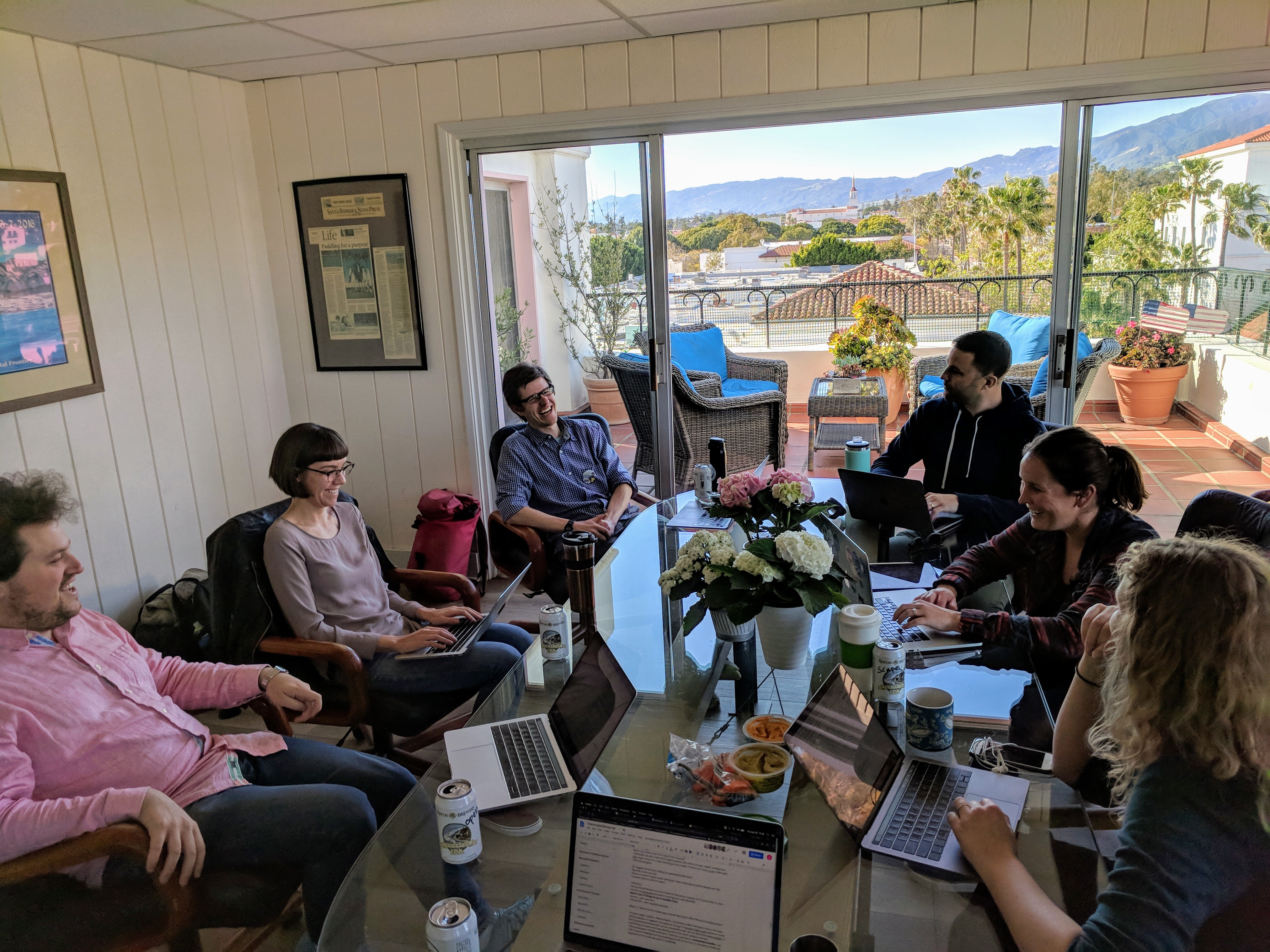 A group of six scientists sitting around a glass table with a sunlit balcony nearby, with laptops out talking and laughing